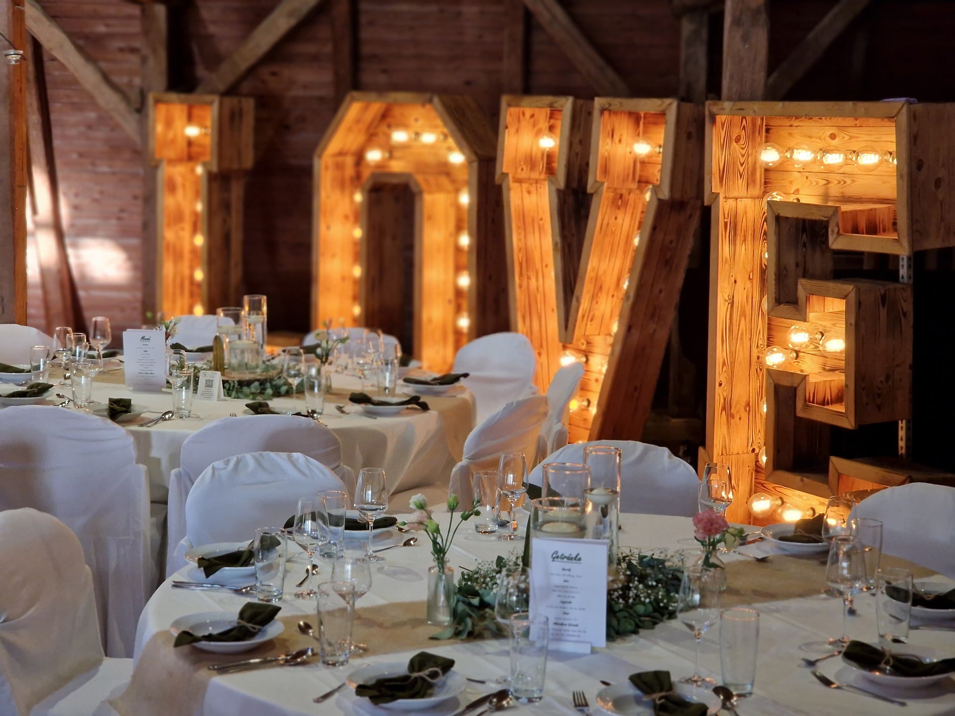 XXL LOVE Letters in geflammter Holzoptik hinter dem Brauttisch in einer Scheune, romantische Hochzeitsdeko mit Vintage-Leuchtmitteln